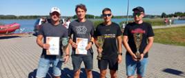 Reprezentacja KM PSP Opole na 3. Mistrzostwach Polski Strażaków PSP w ramach IX edycji Triathlonu Kraśnik – Przegrywają Jedynie Nieobecni. W strażacy w ubraniach cywilnych pozują do zdjęcia trzymając trzy puchary i dwa dyplomy. Funkcjonariusze zdobyli trzecie miejsce w kategorii sztafet PSP. W tle widoczny akwen oraz samochód z pontonem.