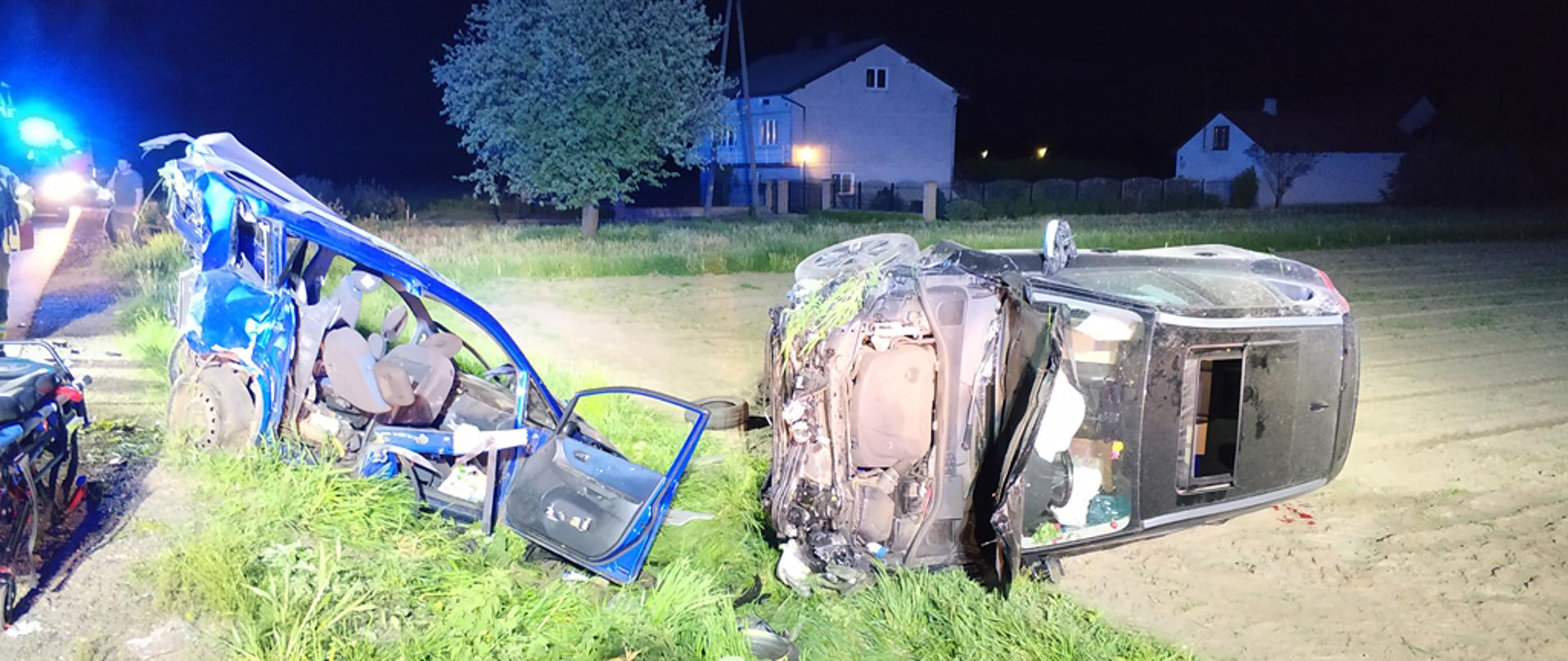 Na zdjęciu widoczne dwa rozbite samochody osobowe. Po lewej stronie koloru niebieskiego, pochylony przodem do przydrożnego rowu, stojący na kołach. Po prawej stronie leży na lewym boku na polu SUV koloru czarnego. Widać także leżące na ziemi, odcięte drzwi z niebieskiego samochodu.