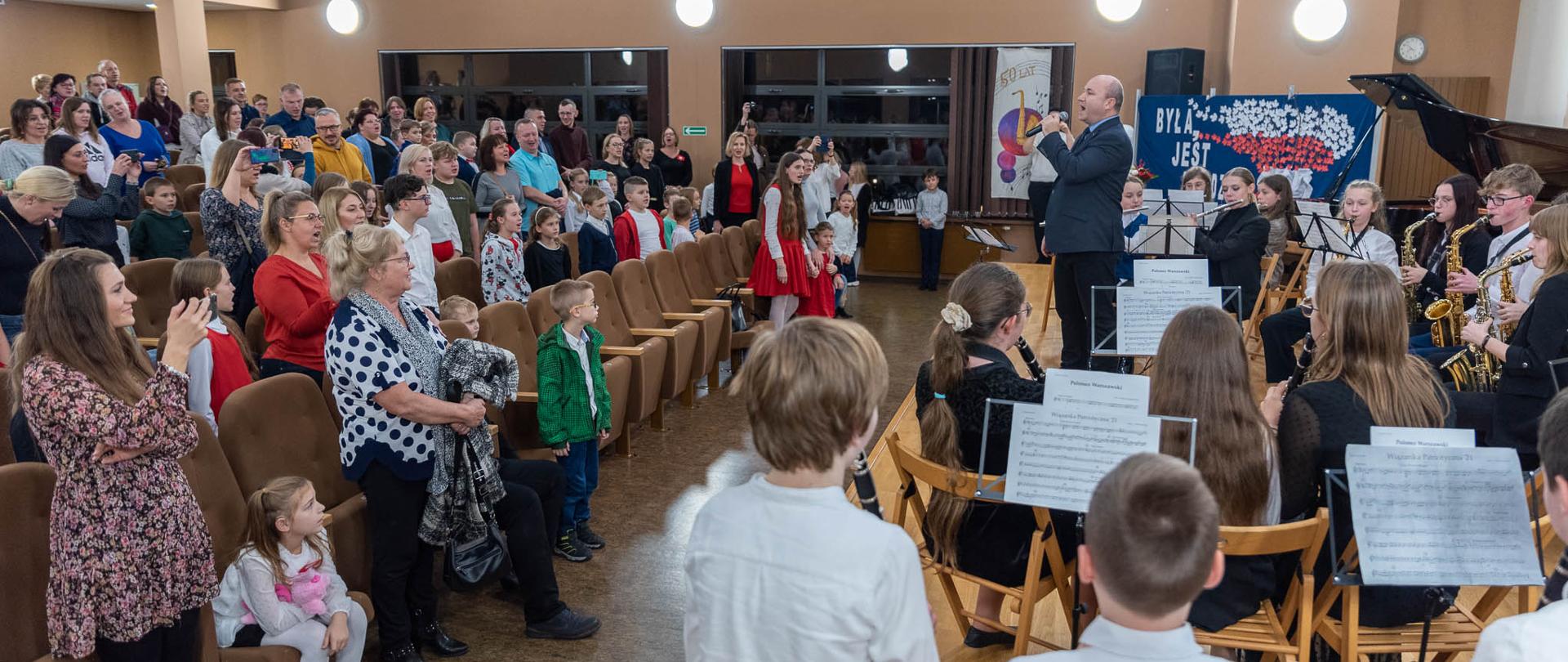 Zdjęcie ze szkolnego koncertu z okazji obchodów Narodowego Święta Niepodległości. Po lewej stronie kadru widnieje stojąca publiczność, która śpiewa pieśń "My, Pierwsza Brygada". Po prawej stronie kadru widnieje dyrygent orkiestry zwrócony do publiczności i śpiewający do mikrofonu. Za nim widać członków orkiestry dętej.