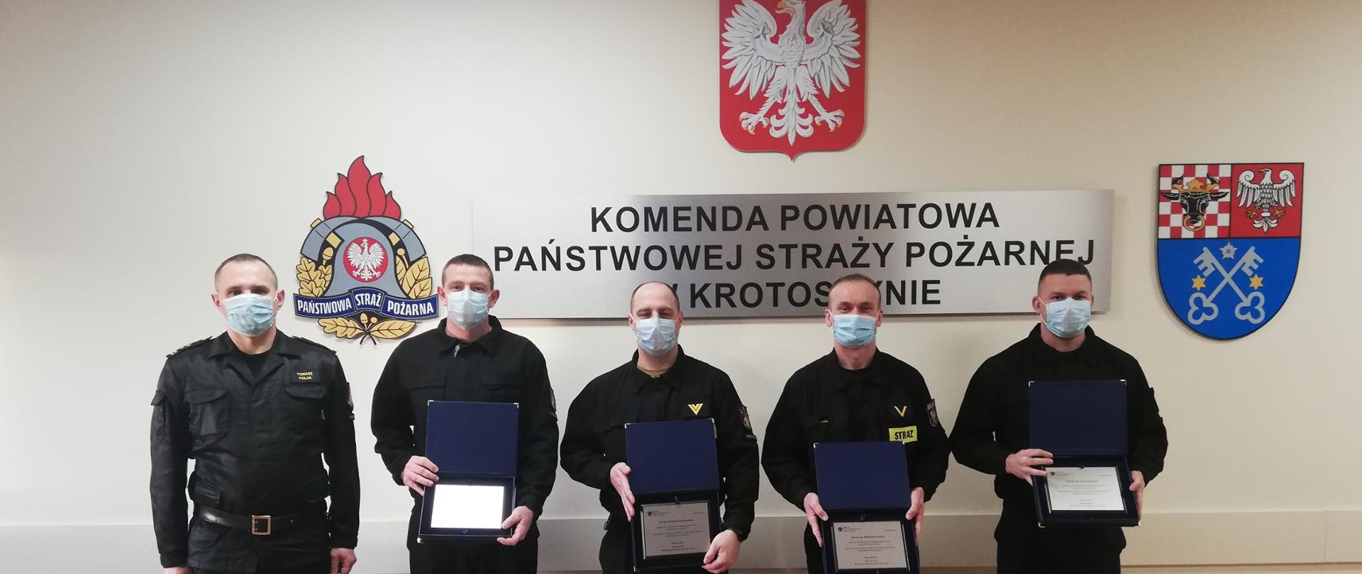 Zdjęcie przedstawia pięciu strażaków Państwowej Straży Pożarnej z Krotoszyna na tle ściany, na której widnieje napis Komenda Powiatowa Państwowej Straży Pożarnej w Krotoszynie, Godło Polski, Herb Powiatu krotoszyńskiego oraz logo PSP. Strażacy w ręku trzymają pamiątkowe ryngrafy. 