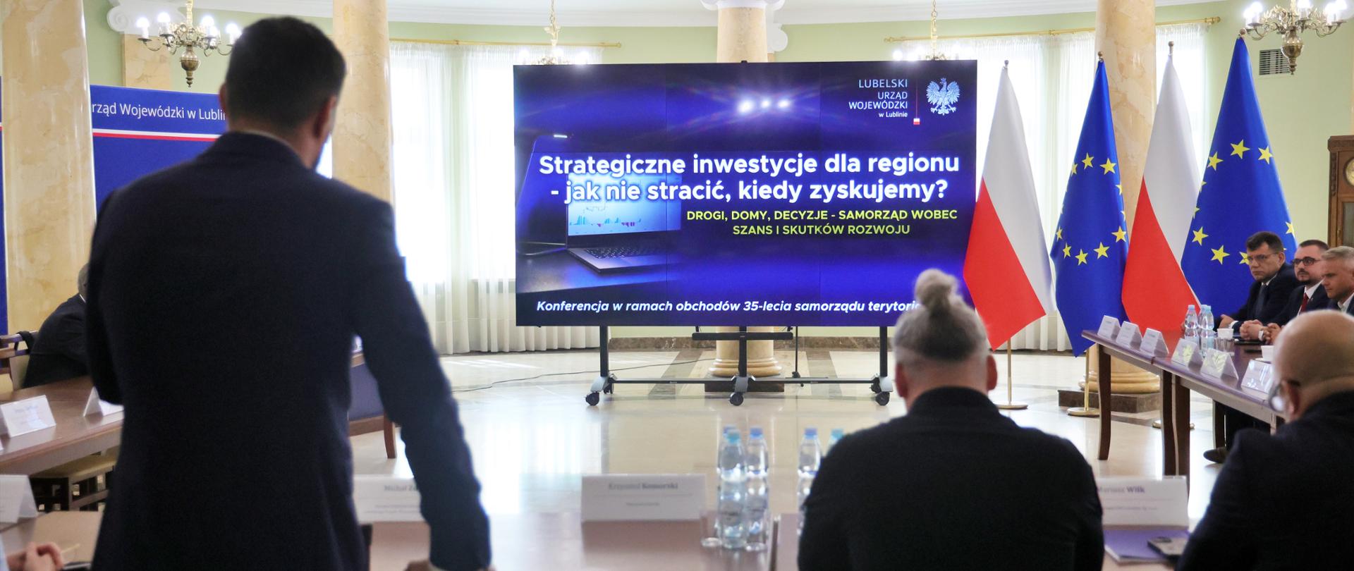 Konferencja "Strategiczne inwestycje dla regionu - jak nie stracić, kiedy zyskujemy?" odbywa się w przestronnej sali Lubelskiego Urzędu Wojewódzkiego, urządzonej w klasycznym stylu z kolumnami. Na środku sali, na dużym ekranie, widoczny jest tytuł konferencji oraz logo LUW i polskiego orła, co podkreśla oficjalny i merytoryczny charakter spotkania. Z przodu, po prawej stronie ekranu, widać polską i unijną flagę, a przy stole prezydialnym siedzą uczestnicy, w tym wojewoda Krzysztof Komorski. Na pierwszym planie, odwrócony tyłem, stoi jeden z prelegentów lub uczestników, co oddaje atmosferę aktywnego udziału w dyskusji.