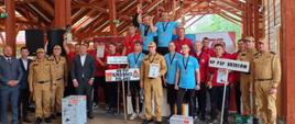 Kolorowa fotografia wykonana w pochmurny dzień podczas wręczenia dyplomów i naród w XXIII Mistrzostwach województwa podkarpackiego w sporcie pożarniczym, Drużyny stoją z dyplomami i nagrodami, w strojach służbowych i dresach pod specjalną drewnianą wiatą.
