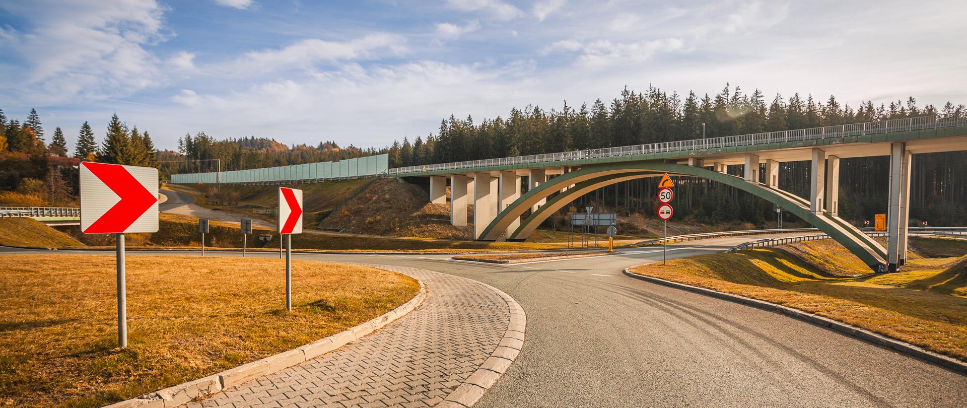 Tarcza ronda o nawierzchni bitumicznej wraz z oznakowaniem pionowym. W tle fotografii droga ekspresowa wraz z mostem w ciągu niej. Obiekt pozwala przekroczyć potok Czerna.