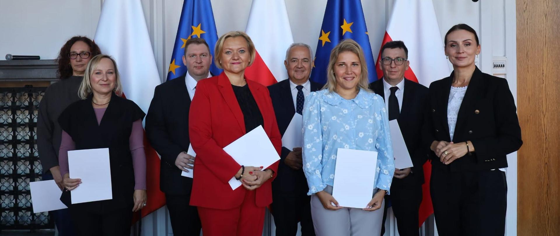 Zdjęcie przedstawia oficjalne zgrupowanie siedmiu kobiet oraz trzech mężczyzn ubranych w eleganckie stroje. Osoby te stoją w rzędzie na tle flag Polski i Unii Europejskiej umieszczonych na stojakach. Niektóre z obecnych osób trzymają w dłoniach białe dokumenty.