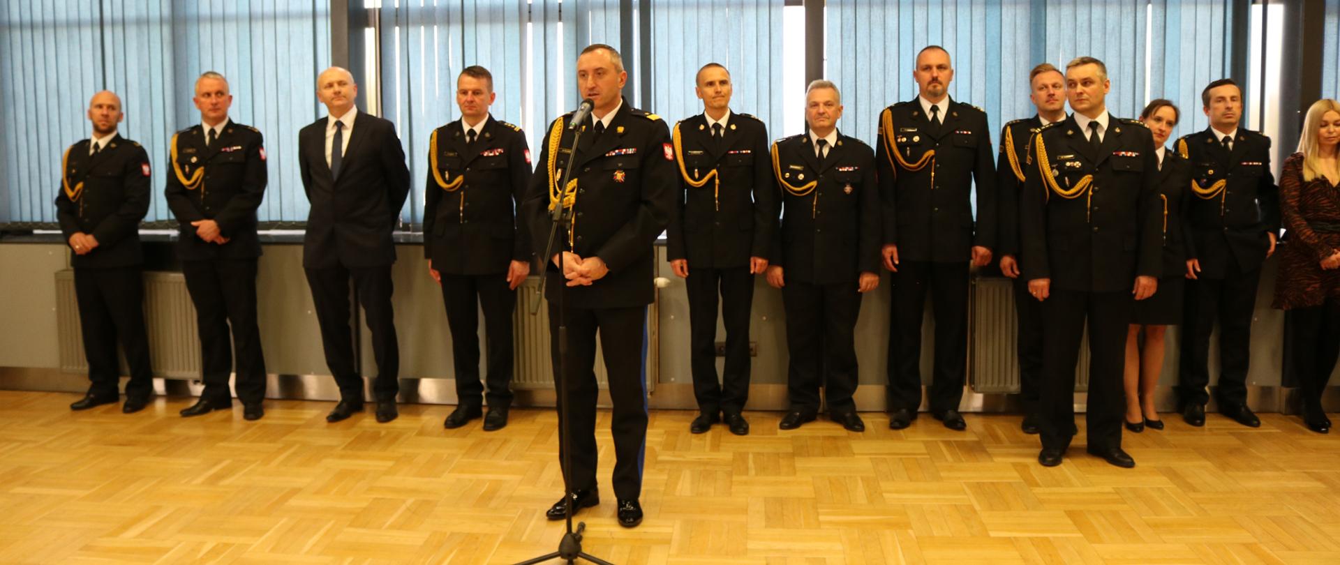 Strażacy ubrani na galowo stoją w rzędzie na uroczystym apelu.