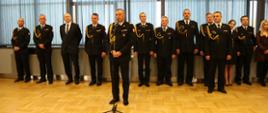 Strażacy ubrani na galowo stoją w rzędzie na uroczystym apelu.