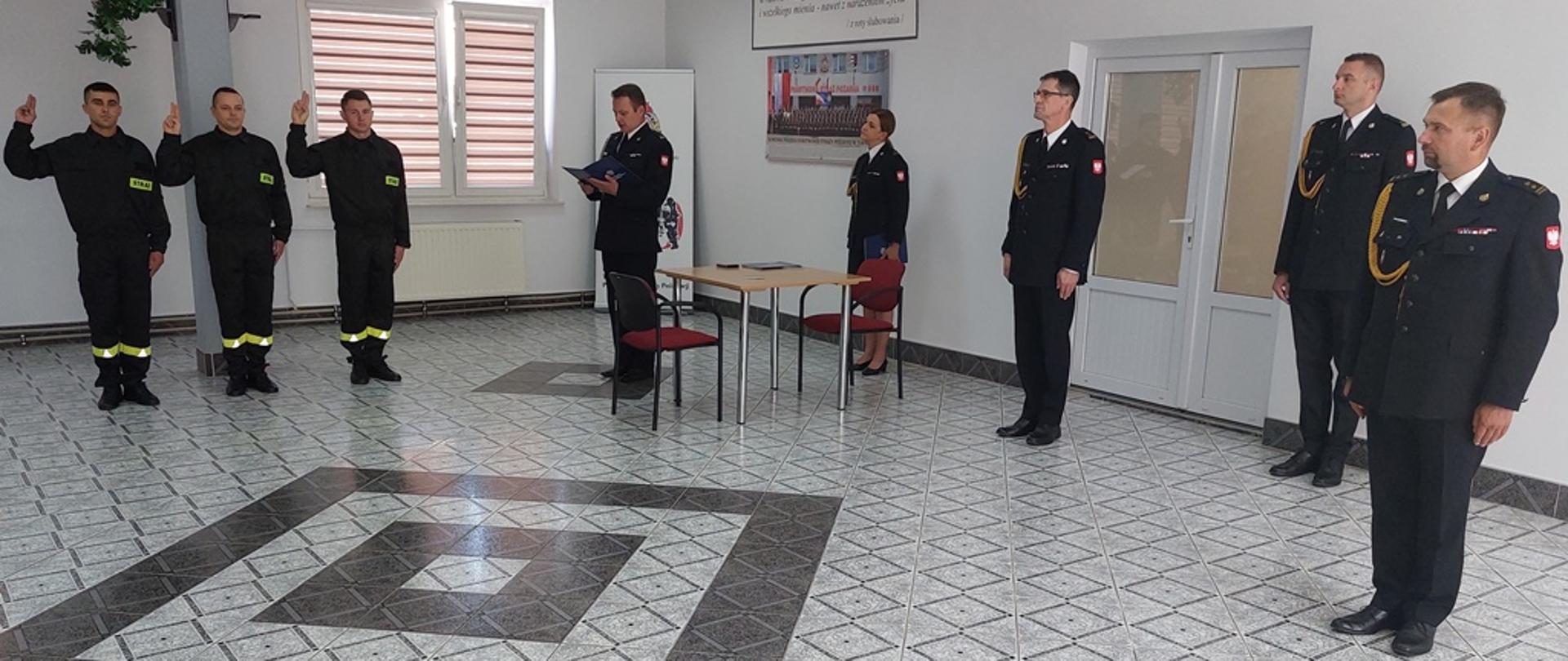 W pomieszczeniu trzech strażaków składa ślubowanie unosząc prawe dłonie. Ślubowanie odbywa się w obecności innych strażaków wchodzących w skład kierownictwa komendy.
