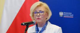Wiceminister Machałek stoi i mówi do mikrofonu na stojaku, za nią niebieska ścianka z napisem Ministerstwo Edukacji i Nauki, obok polska flaga.