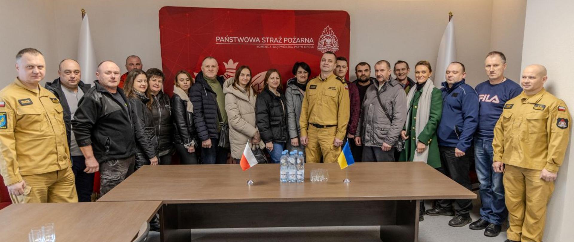 Ukraińscy strażacy ochotnicy i samorządowcy stoją wspólnie z st. bryg. Arkadiuszem Kuśmierskim p.o. Opolskiego Komendanta Wojewódzkiego Państwowej Straży Pożarnej do wspólnego zdjęcia grupowego. Wszyscy znajdują się w sali Ośrodka Szkolenia KW PSP w Opolu. W tle biało czerwone Flagi Rzeczypospolitej Polskiej oraz czerwony banner Państwowej Straży Pożarnej. Na stole stoją małe flagi Polski i Ukrainy. Na zdjęciu ponadto w ubraniu służbowym stoi dwóch oficerów Ośrodka Szkolenia KW PSP w Opolu.