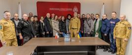 Ukraińscy strażacy ochotnicy i samorządowcy stoją wspólnie z st. bryg. Arkadiuszem Kuśmierskim p.o. Opolskiego Komendanta Wojewódzkiego Państwowej Straży Pożarnej do wspólnego zdjęcia grupowego. Wszyscy znajdują się w sali Ośrodka Szkolenia KW PSP w Opolu. W tle biało czerwone Flagi Rzeczypospolitej Polskiej oraz czerwony banner Państwowej Straży Pożarnej. Na stole stoją małe flagi Polski i Ukrainy. Na zdjęciu ponadto w ubraniu służbowym stoi dwóch oficerów Ośrodka Szkolenia KW PSP w Opolu.