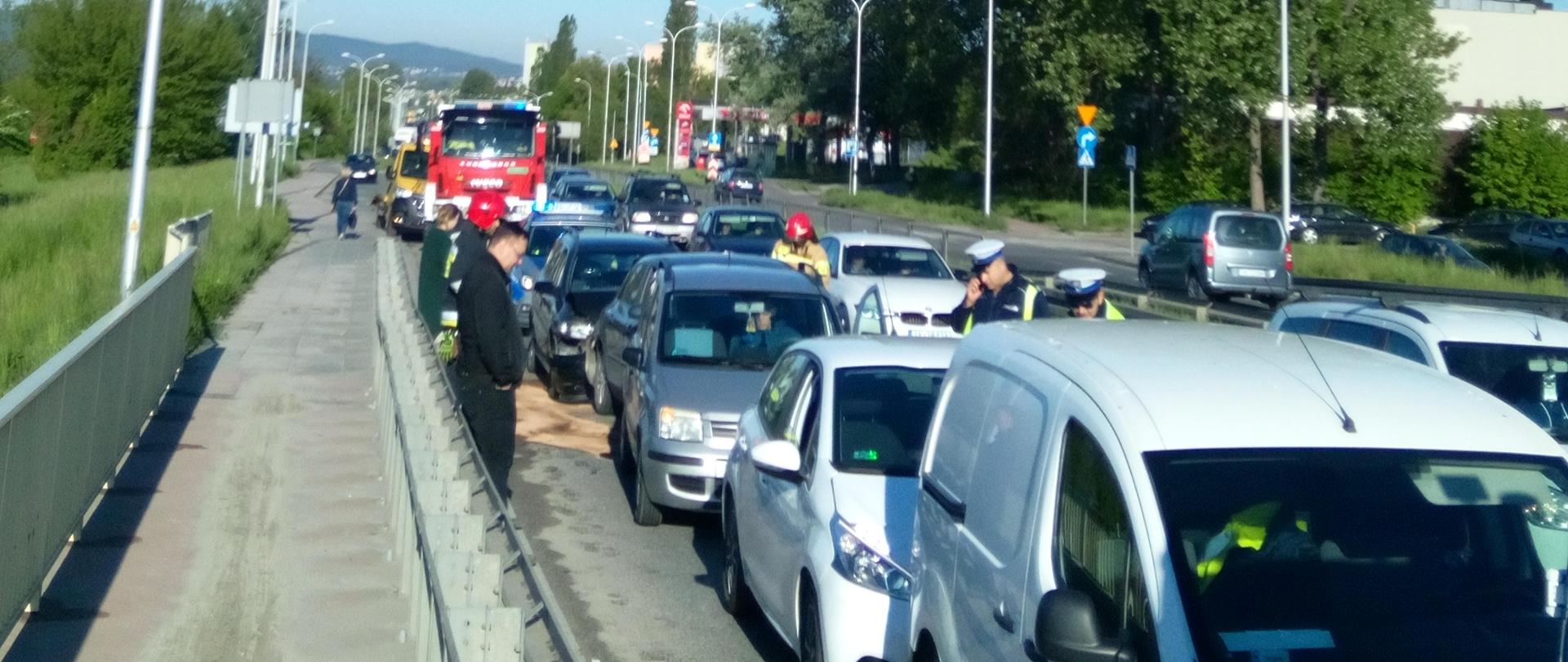 Na zdjęciu widać ulicę na której w rzędzie stoją samochody które brały udział w zderzeniu. W oddali widać służby ratownicze wykonujące swoje czynności na miejscu zdarzenia.