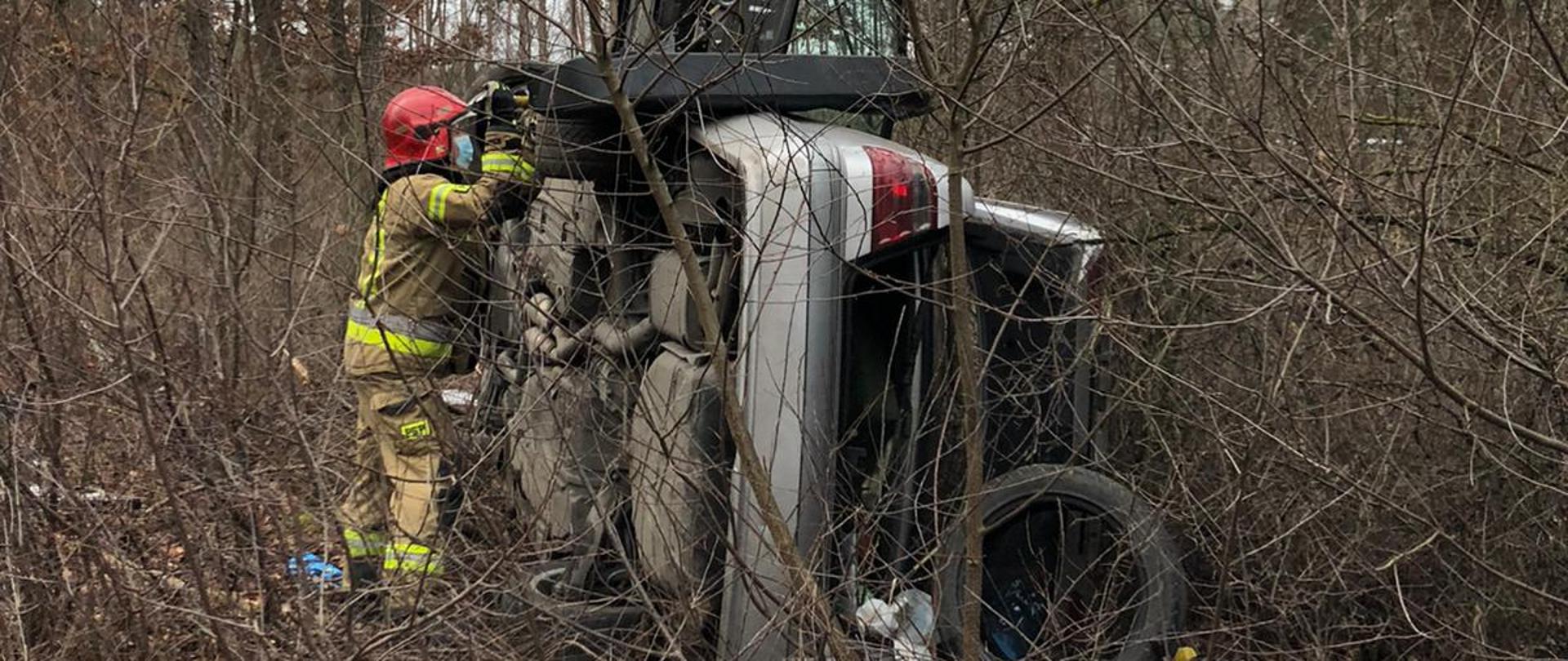 Zdjęcie przedstawia srebrny pojazd marki Mercedes z otwartym bagażnikiem i drzwiami, leżący na lewym boku w zaroślach. Przy samochodzie znajduje się strażak sprawdzający czy nie doszło do wycieków płynów eksploatacyjnych ze samochodu.
