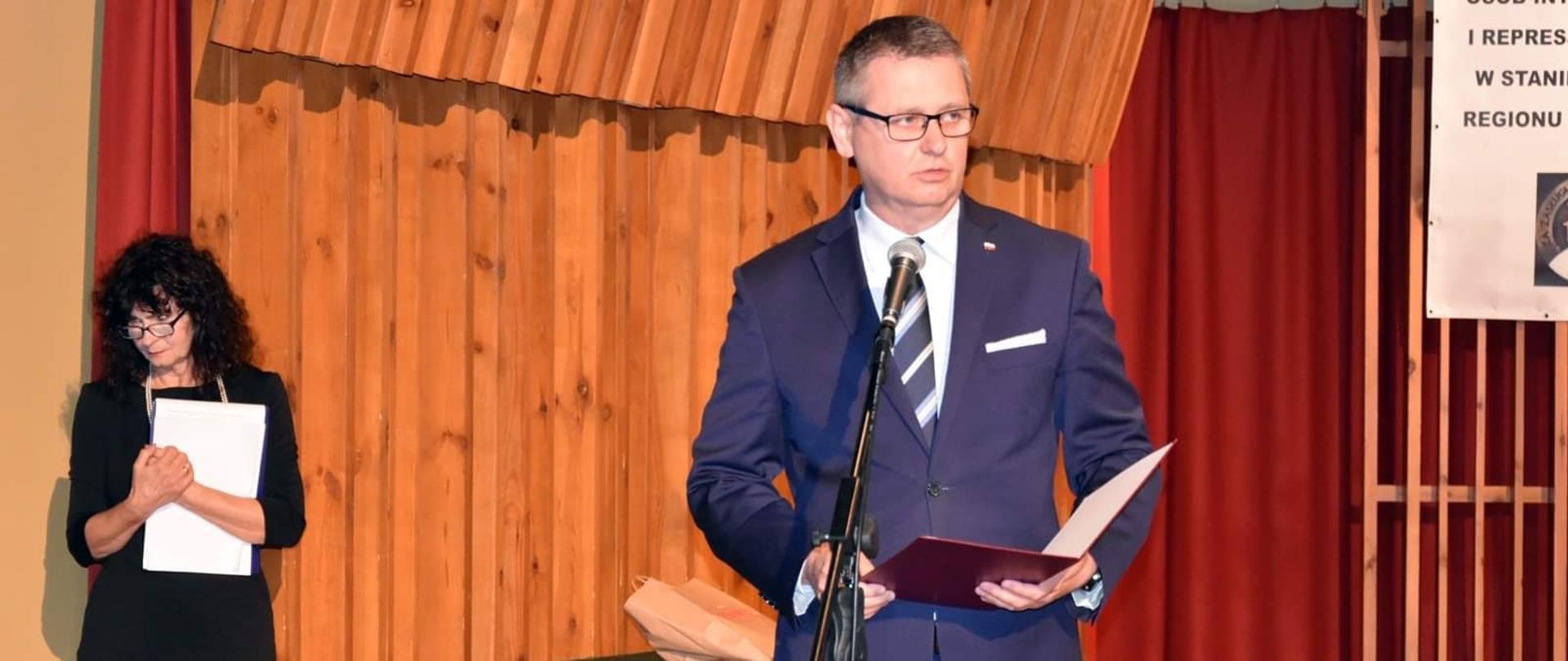 Na pierwszym planie widać elegancko ubranego mężczyznę, który odczytuje list. Na drugim planie elegancko ubrana kobieta wsłuchuje się w słowa listu. 