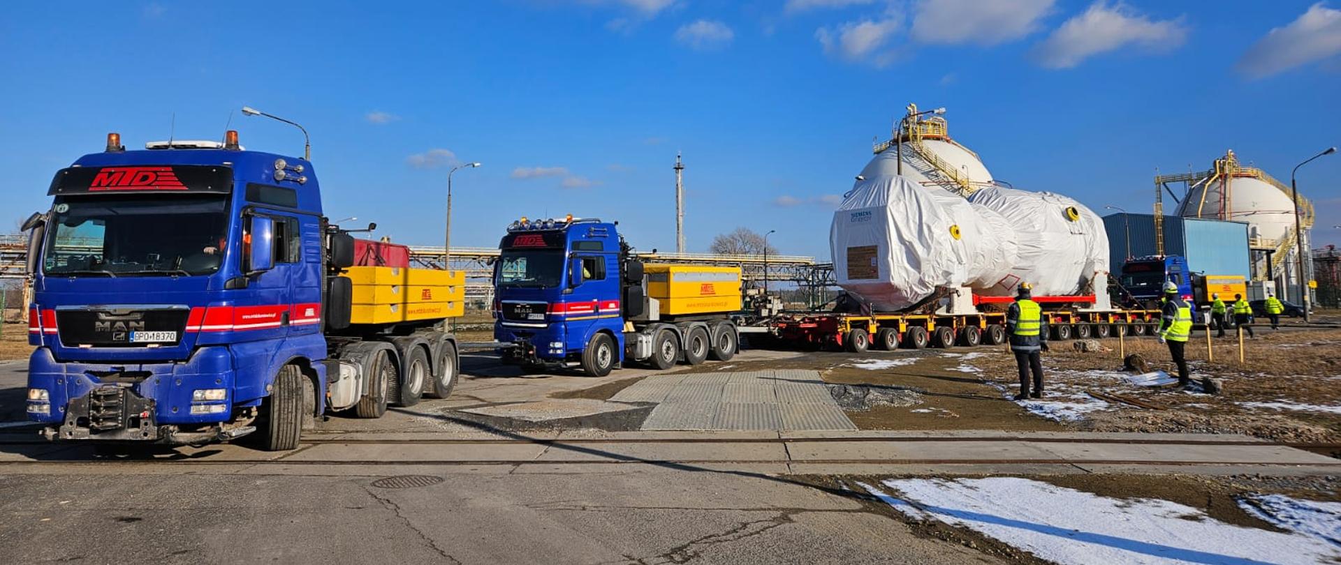 Na zdjęciu widać dwa niebieskie ciężarówki z czerwonymi i żółtymi elementami, oznaczone logo. Ciężarówki przewożą duże, owinięte białą folią ładunki na wieloosiowych naczepach. W tle widać przemysłowe zbiorniki oraz kilka osób w żółtych kamizelkach odblaskowych, które wydają się nadzorować operację transportu. Na ziemi leży trochę śniegu, a niebo jest jasne i niebieskie z kilkoma chmurami.