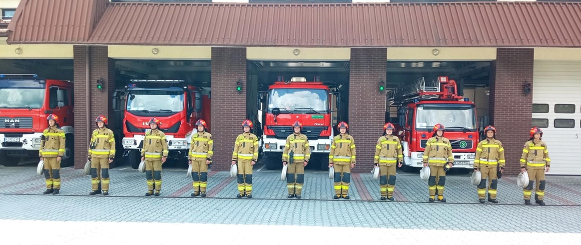 Na zdjęciu widocznych 11 strażaków ubranych w ubrania bojowe czerwone hełmy trzymających w ręku po jednym odcinku węża W -52 przygotowujących się do wykonania zadania w ramach nominacji #UkrainaChallenge. Za ich plecami widoczny budynek Komendy Powiatowej PSP w Gorlicach i otwarte bramy oraz czerwone samochody pożarnicze. 