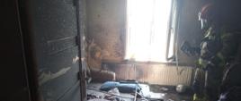 Strażak z kamerą termowizyjną i sprzęcie ochronnym stoi w okopconym i częściowo spalonym pomieszczeniu mieszkalnym.