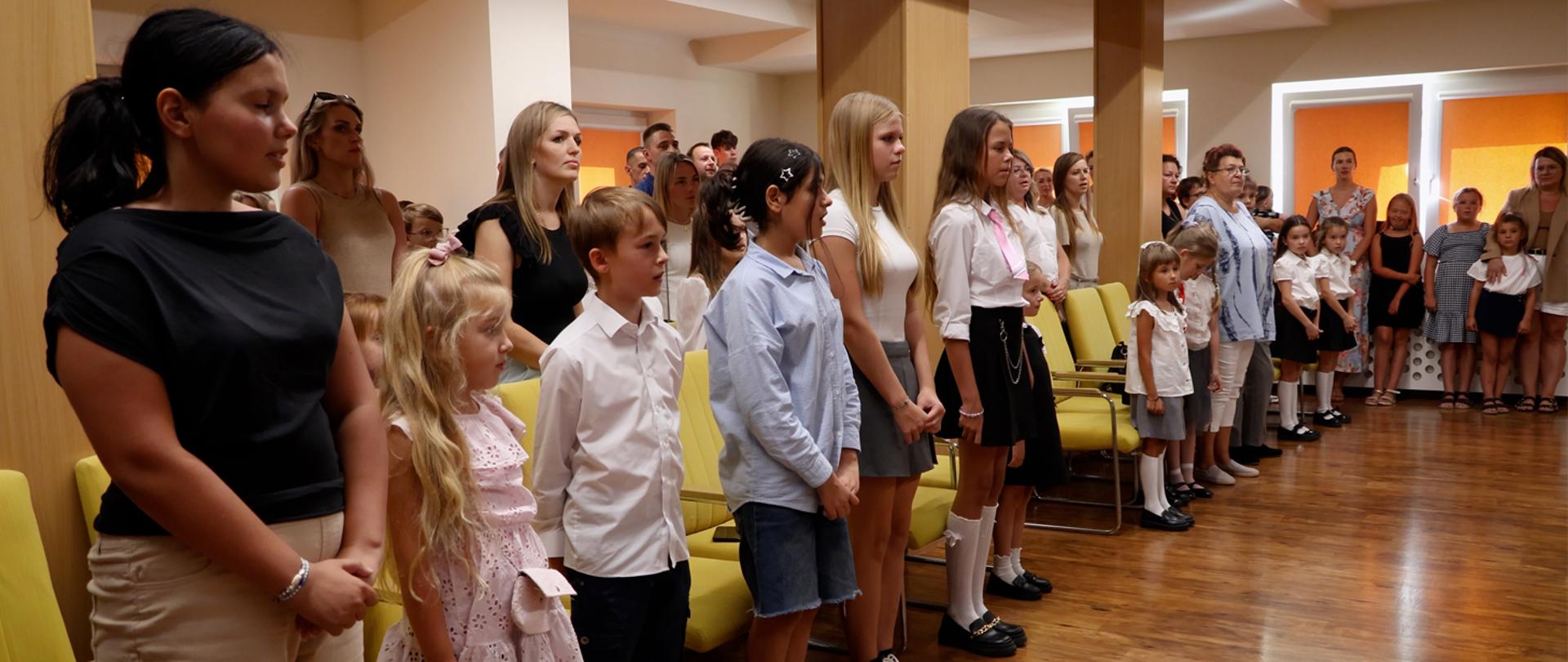 Na zdjęciu duża grupa ludzi, dorosłych i uczniów stoi w auli szkolnej.