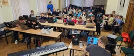 Grupa dzieci i nauczycieli, którzy wzięli udział w warsztatach z produkcji muzycznej. Osoby siedzą przy biurko mając przed sobą laptopy.
