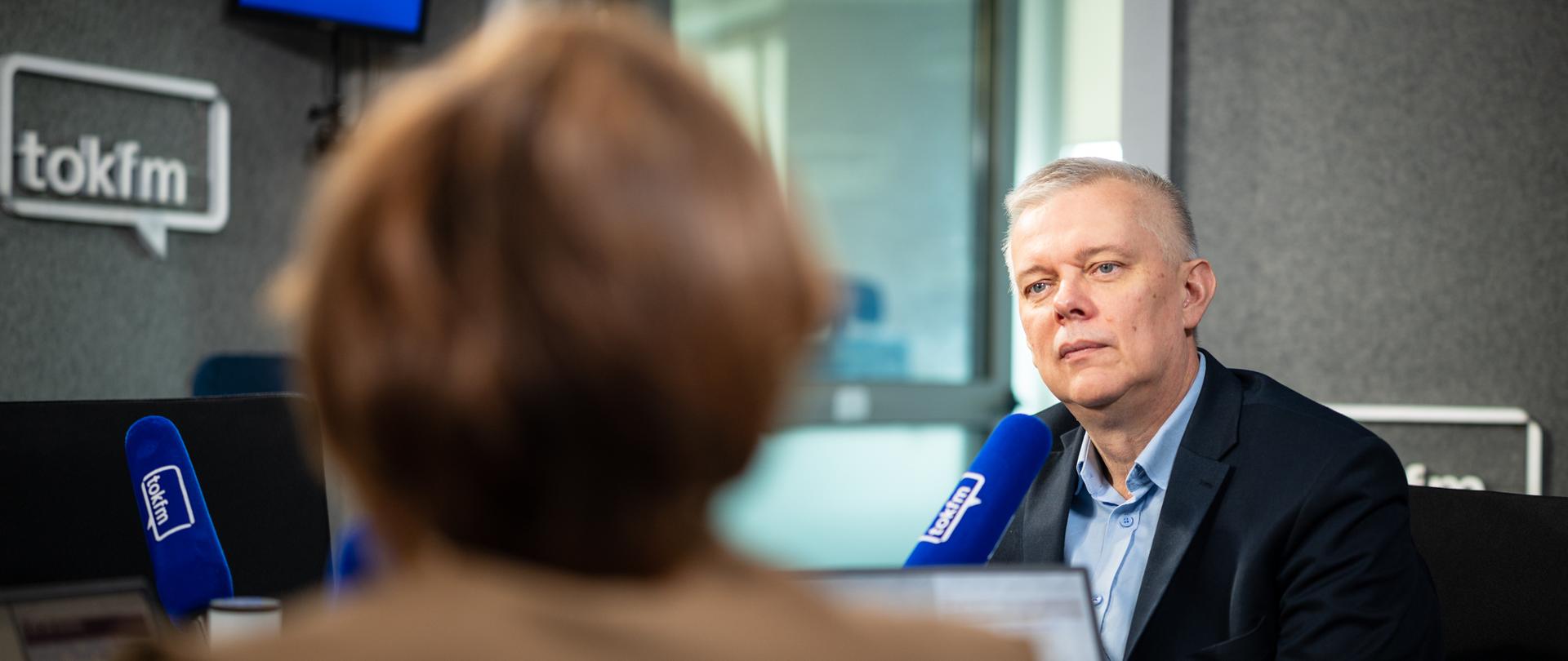Na zdjęciu wnętrze studia radiowego. Przy stole przed niebieskimi mikrofonami TOK FM siedzą - minister Tomasz Siemoniak (po prawej) oraz redaktor Karolina Lewicka (po lewej, tyłem do obiektywu). W tle szyba, szare ściany z białym logo TOK FM.