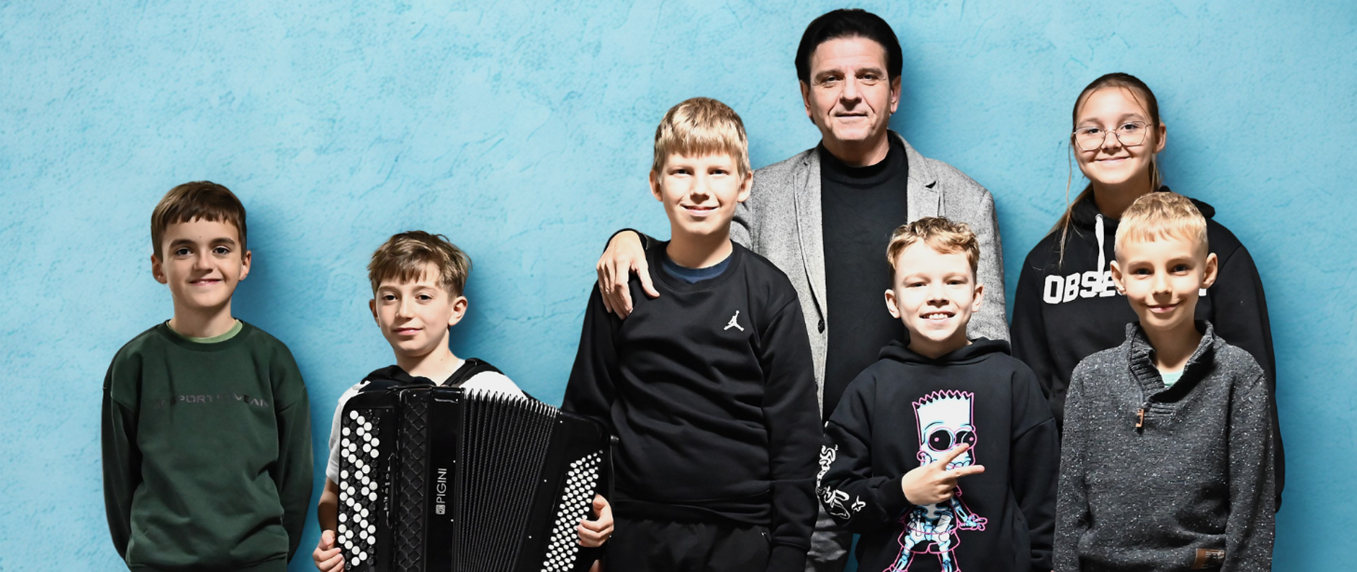 grupka kilku osób, jedna z nich ma założony akordeon, jasnoniebieskie tło