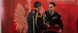Zdjęcie przedstawia oficjalną uroczystość wręczenia wyróżnienia w Państwowej Straży Pożarnej. Dwóch wysokich rangą oficerów w galowych mundurach stoi przed czerwonym banerem z wizerunkiem białego orła i napisem „Państwowa Straż Pożarna”. Jeden z nich przekazuje drugiemu eleganckie czerwone pudełko, prawdopodobnie zawierające odznaczenie lub symboliczny upominek. Oficer odbierający wyróżnienie ma lekki uśmiech na twarzy, co sugeruje podniosłą, ale także serdeczną atmosferę. Na pierwszym planie widoczny jest mikrofon, co wskazuje, że ceremonia odbywa się publicznie, w obecności innych uczestników. Całość podkreśla oficjalny i uroczysty charakter wydarzenia.