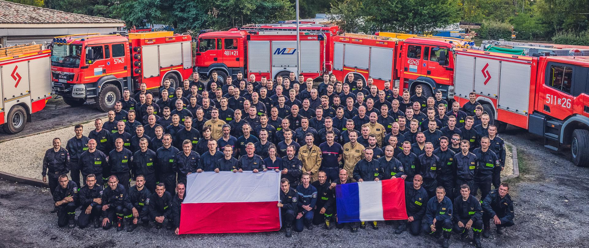 Zdjęcie grupowe modułu GFFFV Poland. Cały stan osobowy pozujący do zdjęcia osoby w pierwszym rzędzie trzymają flagę Francji i Polski. W tle widoczne ustawione w szeregu samochody i maszt flagowy z polską flagą. 