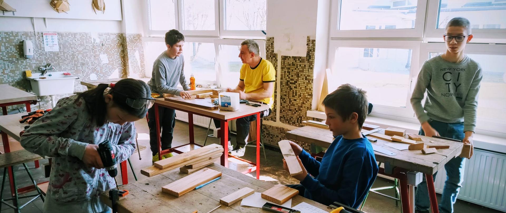 Uczniowie w pracowni technicznej pracują przy stołach warsztatowych nad projektami z drewna.
Na pierwszym planie dziewczynka używa wiertarki, a chłopiec obok przygląda się drewnianemu elementowi. W głębi sali widać kolejnych uczniów oraz nauczyciela w żółtej koszulce. Na stołach leżą narzędzia: piła, kątownik i ołówki, a na ścianie wiszą drewniane modele owadów.
