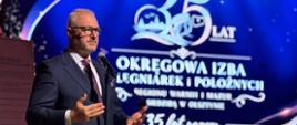 mężczyzna w garniturze, wojewoda, przemawia na scenie, na niebieskim tle