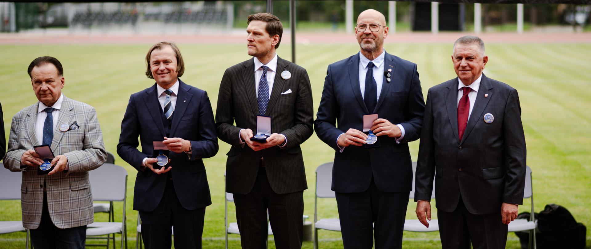 Zdjęcie z otwarcia stadionu w Płońsku. Uczestnicy stoją w rzędzie, prezentując medale. Drugi od prawej minister sportu i turystyki Jakub Rutnicki. W tle murawa stadionu