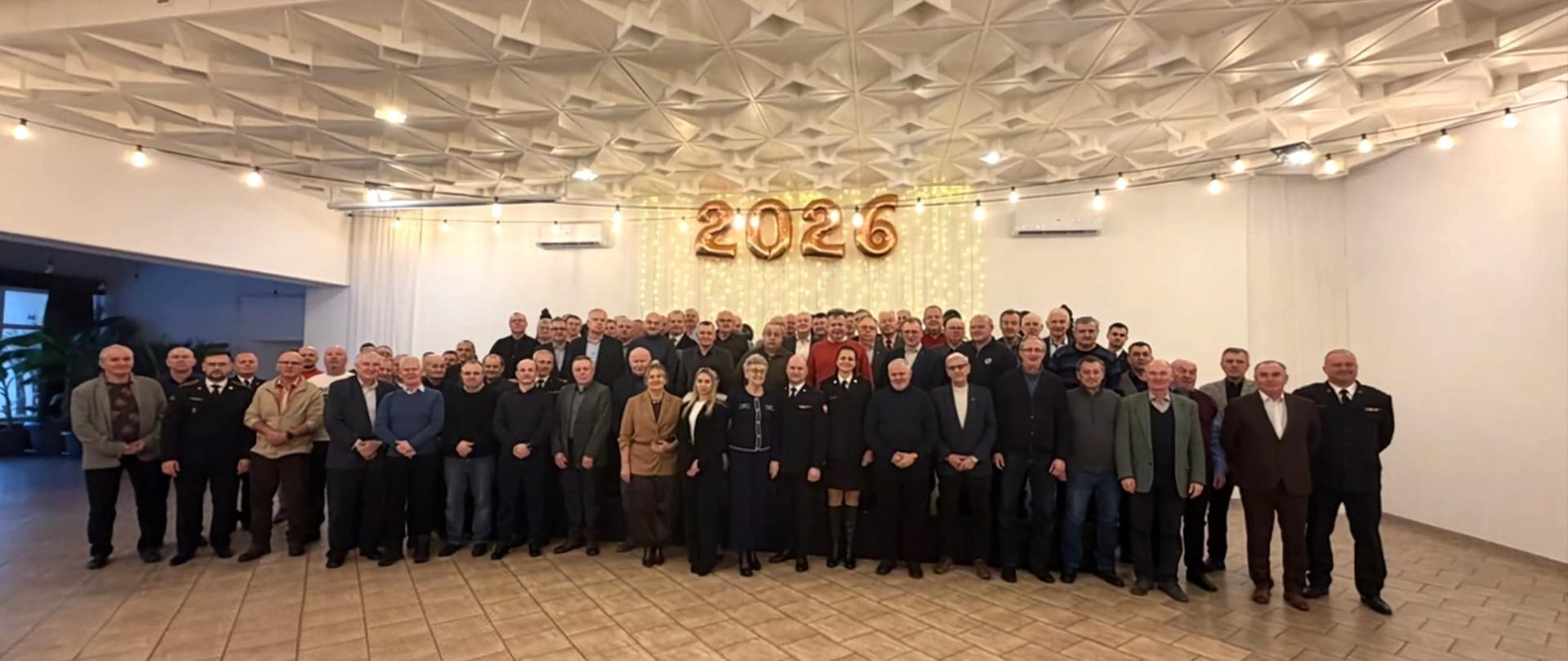 Na zdjęciu widać dużą grupę osób pozujących do wspólnej fotografii w przestronnej sali bankietowej.
Oto szczegóły tego, co znajduje się na obrazku:
Grupa ludzi: W centrum znajduje się kilkadziesiąt osób, w większości mężczyzn, ubranych w stroje wizytowe (garnitury, marynarki) oraz mundury (prawdopodobnie strażackie lub paramilitarne). W centralnej części grupy stoi również kilka kobiet.
Dekoracje: Nad grupą wiszą duże, złote balony tworzące cyfrę "2026". Za ludźmi widoczna jest kurtyna świetlna z drobnych lampek, co sugeruje charakter uroczysty, noworoczny.
Wnętrze: Sala ma charakterystyczny, geometryczny sufit kasetonowy, z którego zwisają sznury z żarówkami dekoracyjnymi. Podłoga wyłożona jest jasnymi płytkami.
Atmosfera: Zdjęcie wygląda na oficjalne podsumowanie wydarzenia.