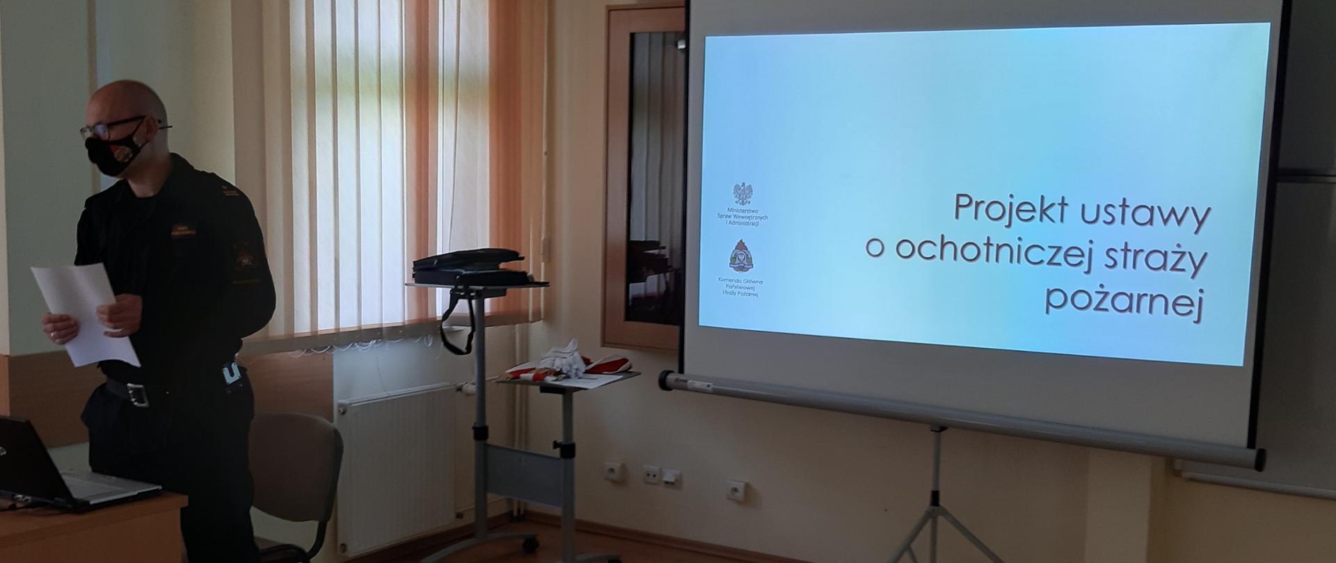 Na świetlicy stoi Komendant Powiatowy PSP, w ręku trzyma dokumenty, za nim wyświetlana jest prezentacja na rzutniku 