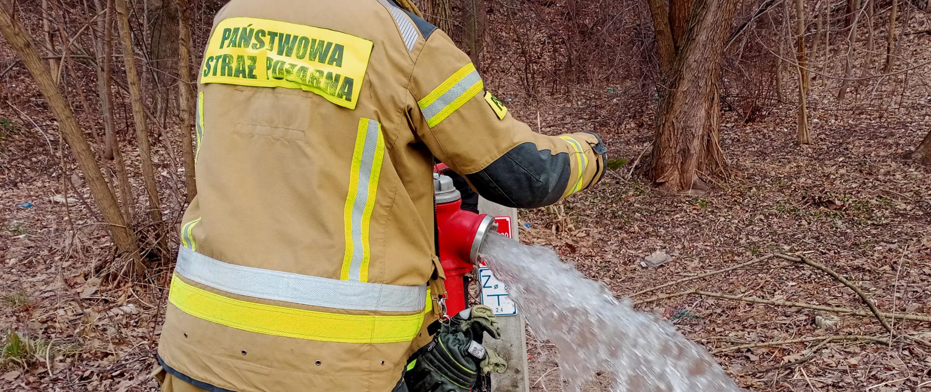 Zdjęcie przedstawia strażaka odkręcającego zawór przy przeciwpożarowym hydrancie zewnętrznym. Z hydrantu wylatuje woda.
