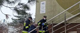 Strażacy wspierają Narodowy Program Szczepień. Na zdjęciu dwóch strażaków w mundurze bojowym pomaga wejść po schodach do budynku przychodni starszej kobiecie.