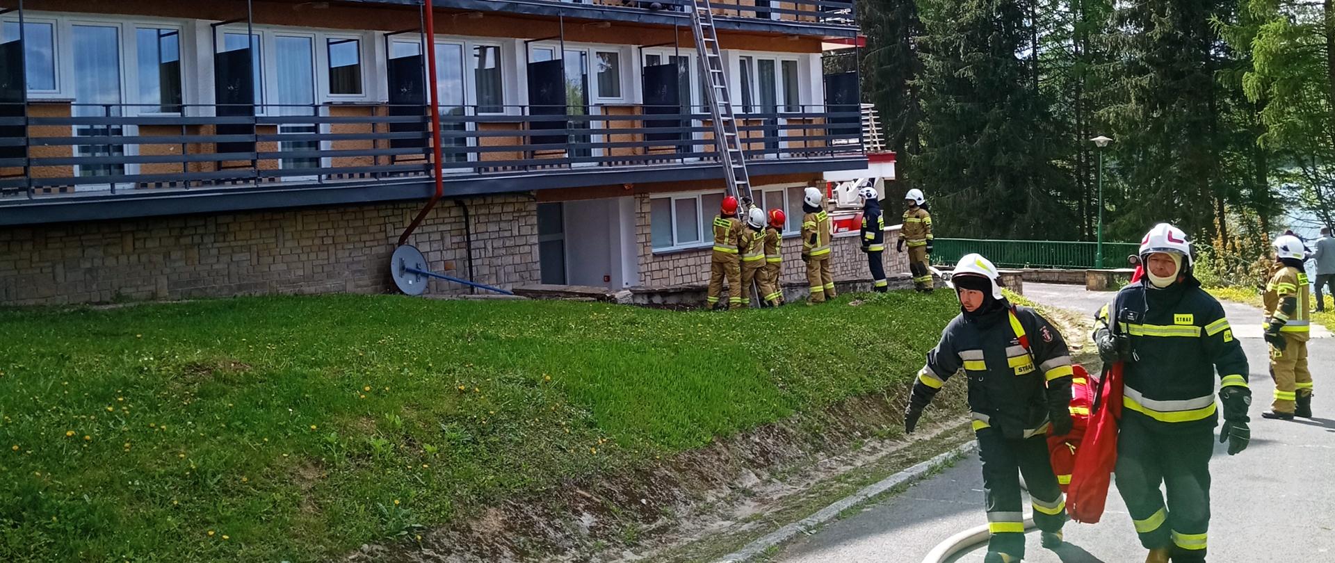 Zdjęcie przedstawia strażaków podczas ćwiczeń z ewakuacji z zadymionego budynku hotelowego. W tle ćwiczących strażaków znajduje się czteropiętrowy budynek hotelowy.