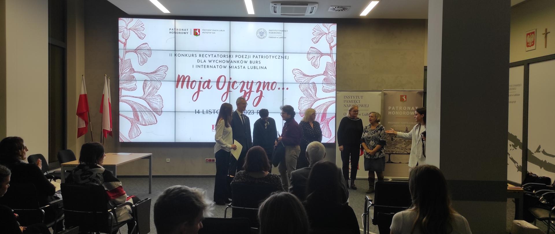 Grupa kilku osób siedzi tyłem do autora zdjęcia patrząc do przodu, gdzie są wręczane dyplomy i nagrody dziewczynie w białej koszuli i chłopcu w koszuli bordowej, w tle na białym podzielonym na kwadraty ekranie wyświetlany napis "II KONKURS RECYTATORSKI POEZJI PATRIOTYCZNEJ DLA WYCHOWANKOW BURS I INTERNATÓW MIASTA LUBLINA Moja Ojczyzno" oraz czerwone motywy roślinne. Po lewej na stojaku stoją trzy biało- czerwone flagi.