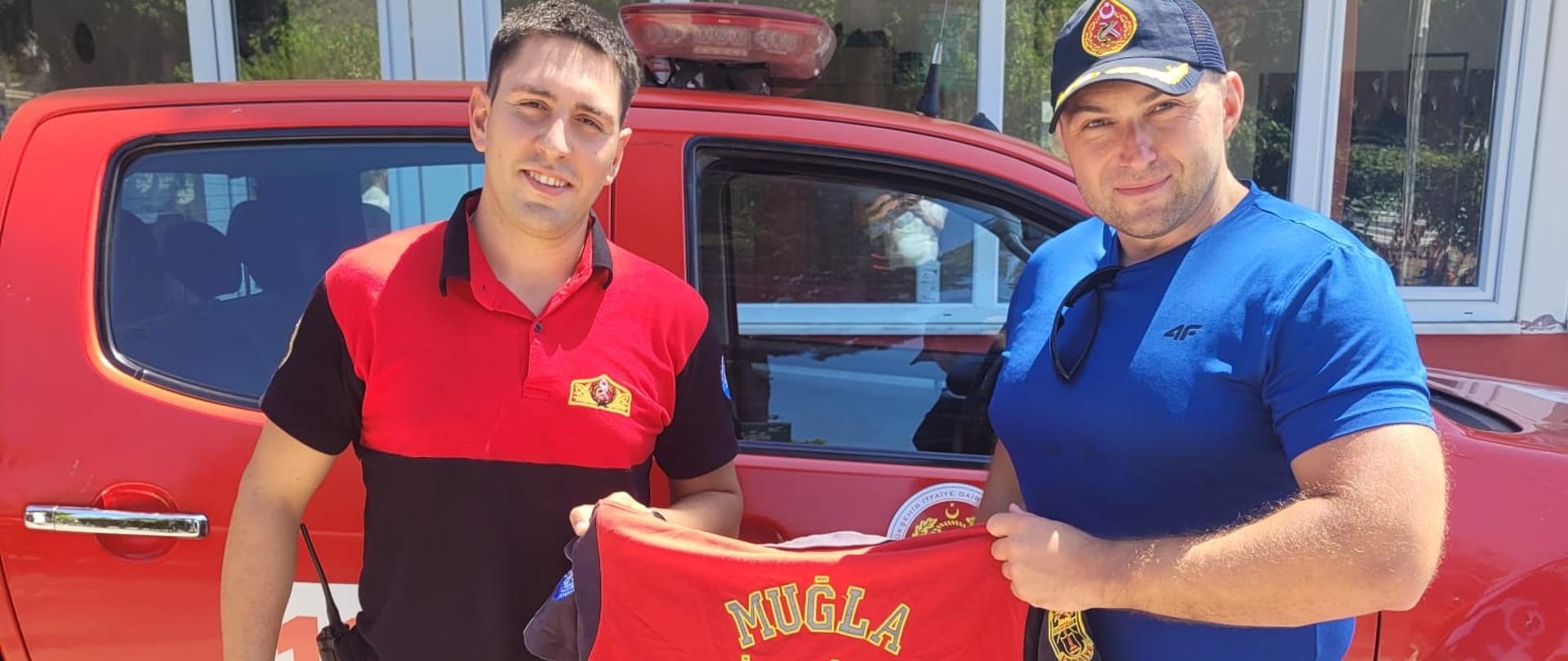 Funkcjonariusz CS PSP w stroju cywilnym oraz funkcjonariusz tureckiej straży pożarnej trzymający wspólnie T-Shirt tureckiej straży pożarnej