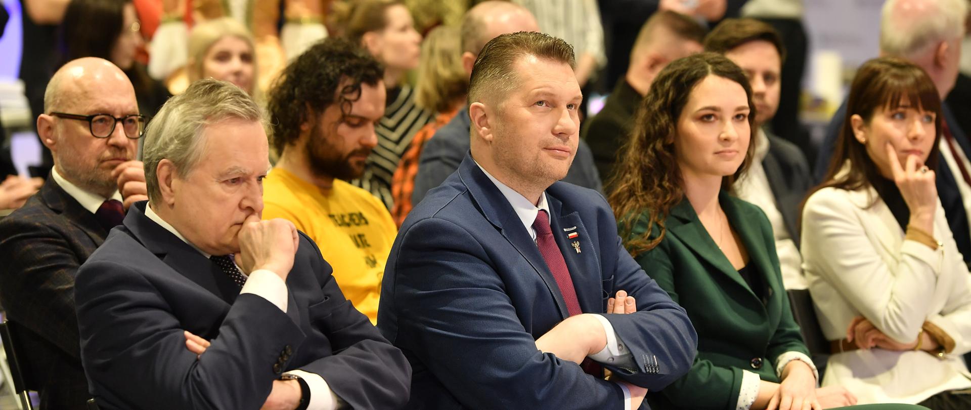 Minister Czarnek i inne osoby siedzą na ustawionych w rząd krzesłach.