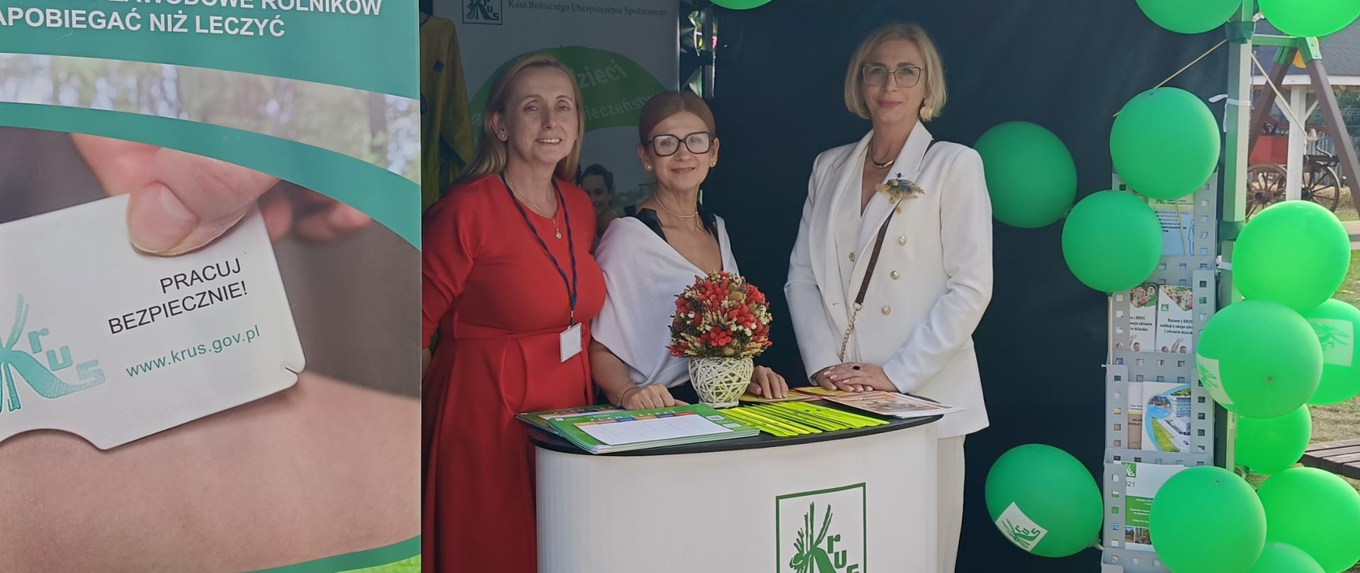 Przy stoisku informacyjno-prewencyjnym Kasy stoją trzy kobiety. Na stoliku przy stoisku znajdują się materiały prewencyjne Kasy (odblaski, plany lekcji oraz ulotki). Stoisko jest udekorowane balonami oraz roll-upami.