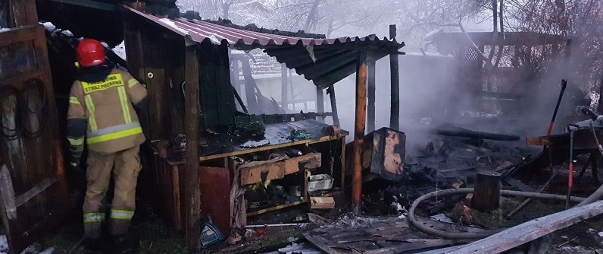 Zdjęcie przedstawia spalony obiekt gospodarczy częściowo kryty blachą falistą koloru czerwonego. Z prawej strony widoczne zgliszcza konstrukcji ścian i dachu zwalone na ziemię i płaty blachy dachowej. Z lewej strony zdjęcia, drzwi wejściowe do obiektu, a w nich obrócony plecami do oglądającego strażak w żółtym ubraniu specjalnym.