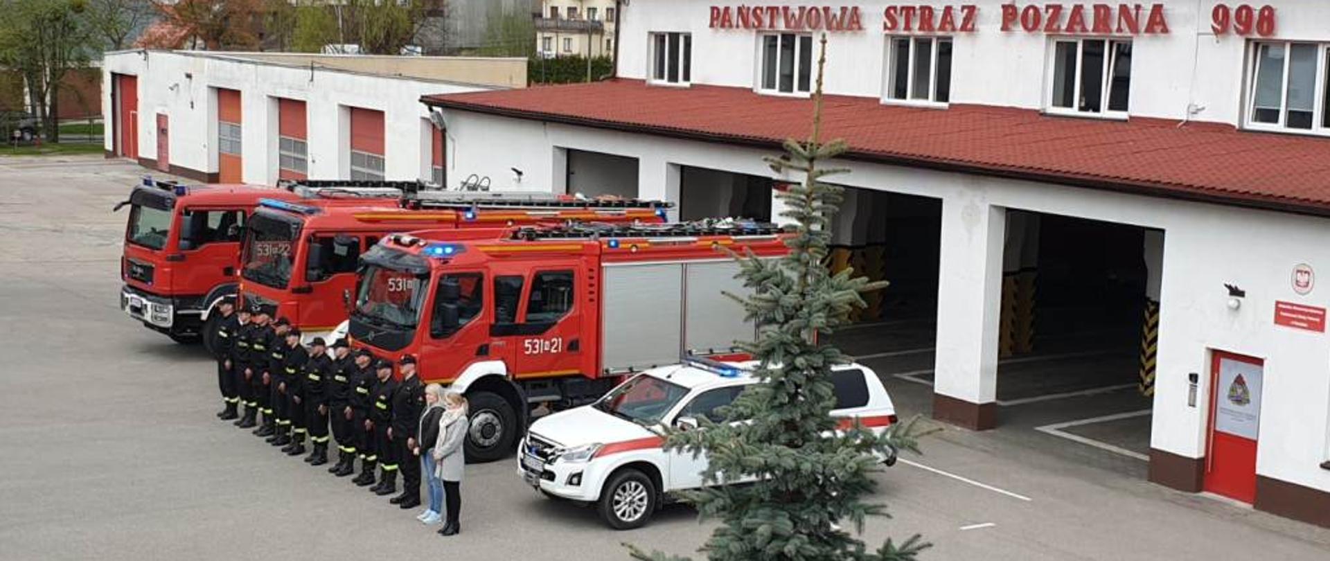 Strażacy i pracownicy cywilni ostródzkiej Komendy Powiatowej PSP stoją na zbiórce przed strażackimi samochodami, w których włączone są sygnały świetlne i dźwiękowe. 