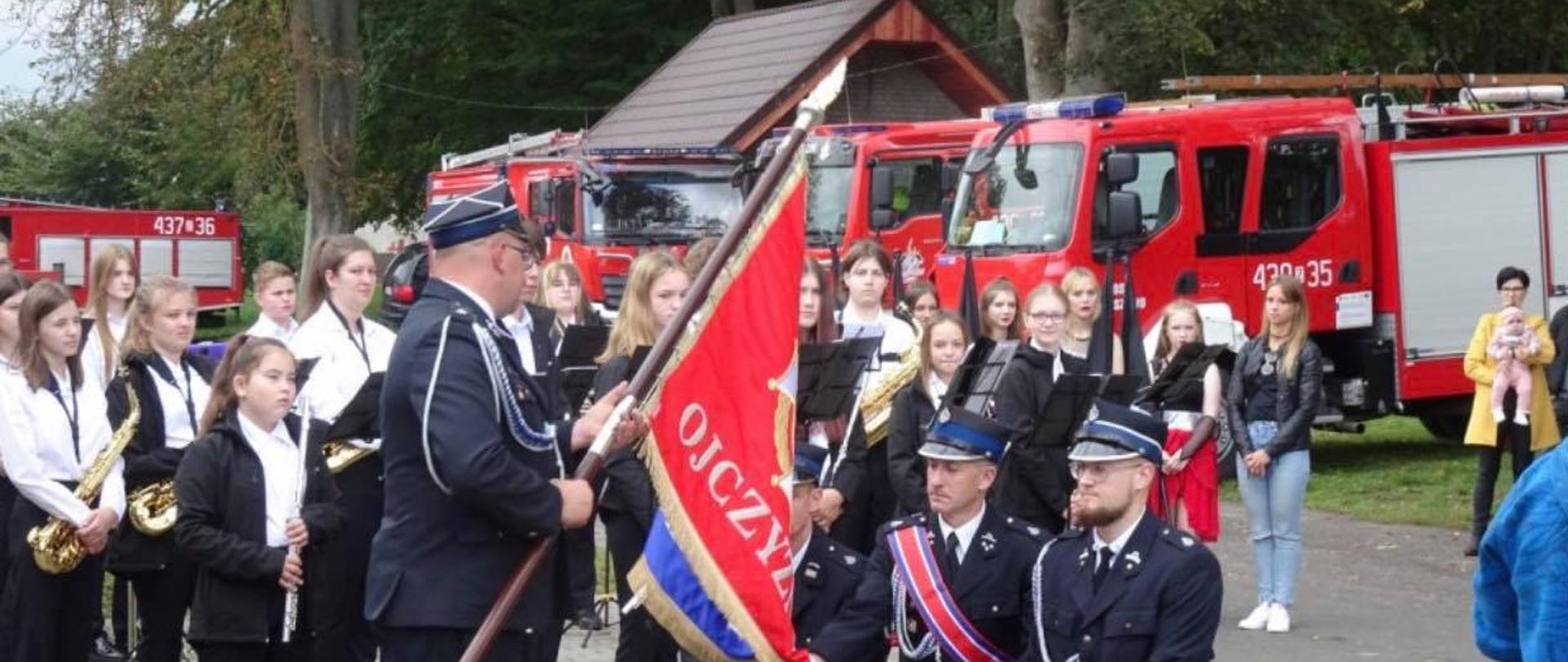 Na zdjęciu poczet sztandarowy któremu wręczany jest sztanadar Ochotniczej Straży Pożarnej. W tle orkiestra oraz samochody ratowniczo-gaśnicze.