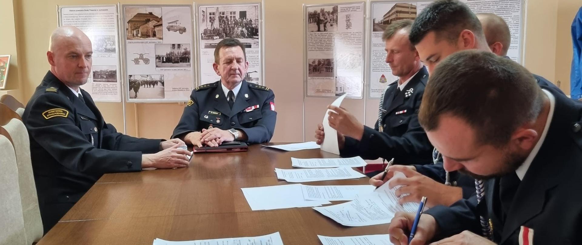 Zdjęcie przedstawia p.o. zastępcy świętokrzyskiego komendanta wojewódzkiego PSP w Kielcach st. bryg. Sławomira Karwata, komendanta powiatowego PSP w Jędrzejowie mł. bryg. Mariusza Stolarczyka oraz druhów jednostek OSP, którzy podpisują umowy na pełnienie funkcji kierowcy w danej ochotniczej straży pożarnej. 