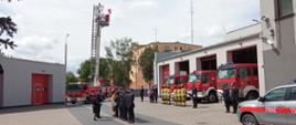 Po lewej jest podnośnik straży pożarnej na którym powiewa flaga państwowa, na środku jest sztandar i strażacy podczas uroczystego apelu, za nimi trzy czerwone samochody straży i budynek komendy