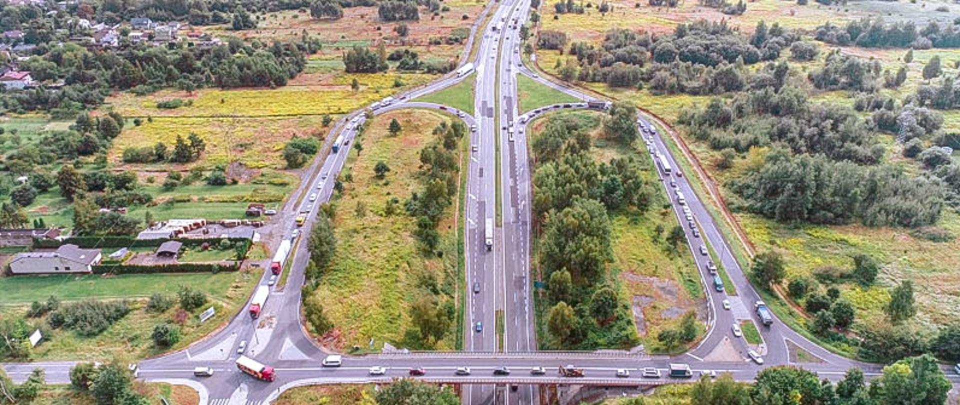 Zdjęcie przedstawia widok z lotu drona na węzeł drogowy na drodze ekspresowej S1. Widoczne poruszające się samochody, w tle mała miejscowość i zadrzewienia.