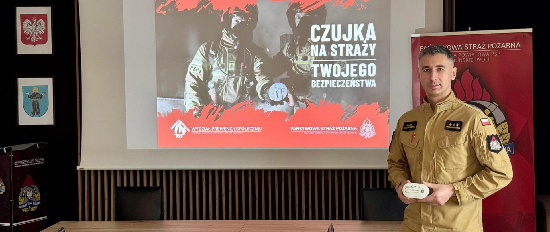 Strażak w piaskowym mundurze stoi obok ekranu z plakatem kampanii „Czujka na straży Twojego bezpieczeństwa”. W ręku trzyma białą czujkę dymu.