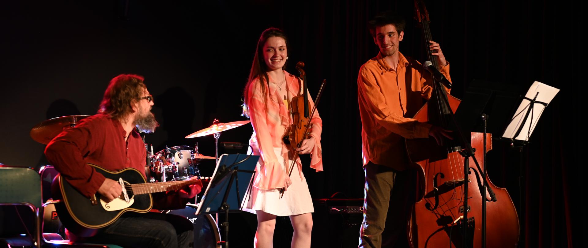 Na oświetlonej scenie są trzy osoby. Pośrodku stoi dziewczyna ze skrzypcami, po prawej stronie chłopak z kontrabasem, a po lewej mężczyzna z gitarą siedzi na krześle. Wszyscy uśmiechnięci, zwróceni w swoją stronę