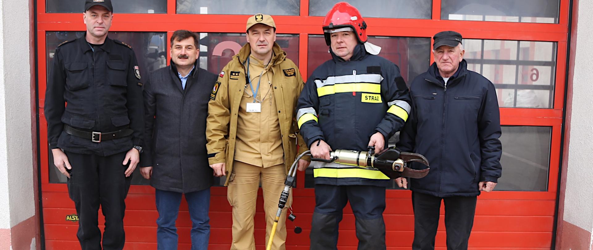 Komendant CS PSP w towarzystwie trzech przedstawicieli OSP oraz naczelnika wydziału technicznego CS PSP stojący w rzędzie na tle bramy garażowej JRG. Przed nimi na macie leżący przekazywany zestaw sprzętu hydraulicznego