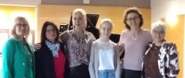 Na zdjęciu widzimy pięć osób – cztery kobiety dorosłe( nauczycieli wraz z prowadzącą warsztaty) oraz jedną nastoletnią dziewczynę ( uczennice)– stojących razem w rzędzie, pozujących do wspólnej fotografii w jasnej sali z drewnianą podłogą. W tle widoczny jest fortepian, co sugeruje, że zdjęcie wykonano w sali muzycznej, po zakończonych warsztatach.Opis postaci od lewej: Pierwsza kobieta po lewej ma jasne, blond włosy, ubrana jest w zieloną marynarkę i ciemne spodnie. Stoi pewnie i uśmiecha się delikatnie. Druga kobieta ma ciemniejsze włosy, okulary, ubrana jest w różowy t-shirt, czarną marynarkę i niebieskie dżinsy. Ma szalik w odcieniach błękitu. W środku stoi młoda dziewczyna – najpewniej uczennica. Ma jasne blond włosy związane do tyłu i nosi jasną bluzkę oraz błękitne, szerokie dżinsy. Trzyma w rękach kartki z nutami.
Czwarta kobieta ma blond włosy spięte do tyłu, ubrana jest w wzorzystą, jasną bluzkę i czarne, skórzane spodnie. Stoi blisko dziewczyny i również się uśmiecha. Ostatnia osoba z prawej ma ciemniejsze, kręcone włosy i ubrana jest w połyskującą jasnoróżową bluzkę z szerokimi rękawami oraz czarne spodnie i szpilki. Ma pogodny wyraz twarzy. Wszyscy stoją blisko siebie, uśmiechają się i patrzą w stronę aparatu. Zdjęcie emanuje pozytywną atmosferą – widać, że to moment dumy i radości po wspólnym wydarzeniu muzycznym.