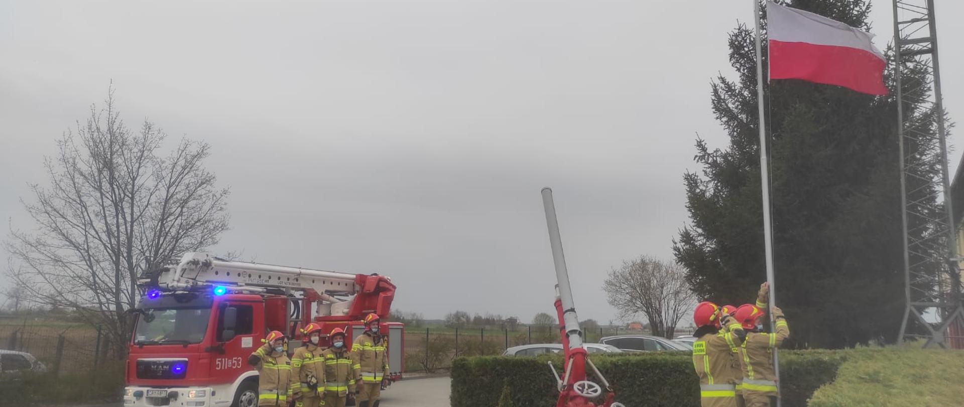 Zdjęcie przedstawia strażaków PSP, którzy z okazji Dnia Flagi Rzeczypospolitej Polskiej podnoszą flagę Polski na maszt. Na zdjęciu po lewej stronie widoczny jest samochód pożarniczy - podnośnik hydrauliczny z włączonymi niebieskimi światłami alarmowymi. Przed pojazdem stoi 4 strażaków, pierwszy z nich salutuje. W środkowej części zdjęcia widać działko wodno - pianowe. W prawej części zdjęcia znajduje się 3 strażaków, którzy stoją przed maszem i podnoszą flagę Polski na maszt.