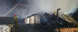 Zdjęci przedstawia pożar budynki inwentarskiego. Na pierwszym planie zdjęcia widać strażaka stojącego na dachu budynku. Na drugim planie widać powalony dach budynku który uległa spaleniu. W oddali widać pracujący podnośnik straży pożarnej.